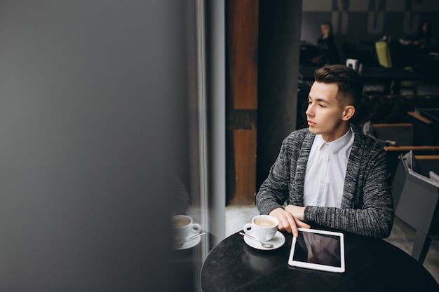 A young professional sitting in a coffee shop, looking attentively at a mentor figure who is pointing at notes on a tablet. The atmosphere is relaxed and informal.
