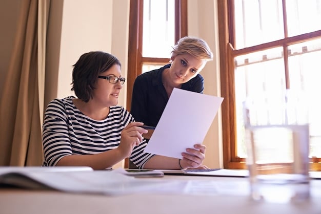 Two people sitting at a desk in a modern office, reviewing documents together. One person is pointing to something on the document, seemingly explaining it to the other. The atmosphere is collaborative and focused.