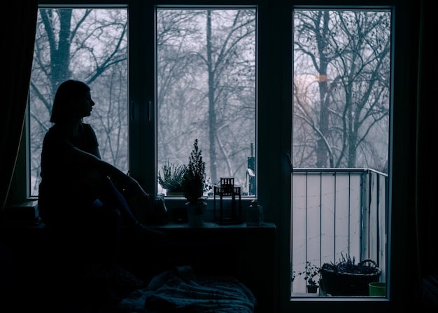 A person sitting alone in a dark room, looking out a window with a pensive expression. Rain streaks the glass, symbolizing sadness and uncertainty.