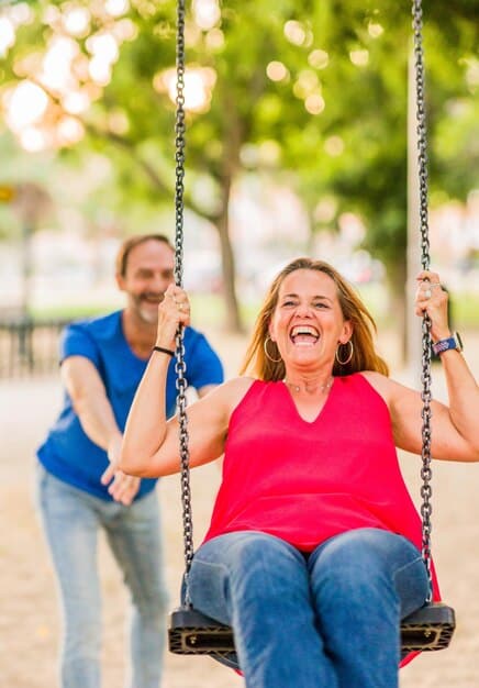 An adult couple holding hands swings laughing at a park. Colorful swings are moving while sunlight covers all over the place.