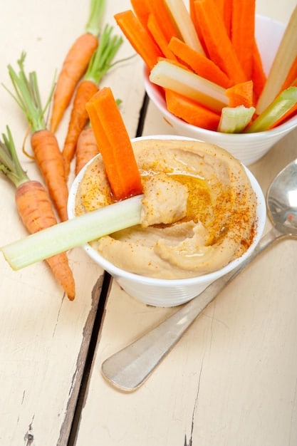 A close-up shot of colorful veggie sticks (carrots, celery, cucumber) arranged attractively around a bowl of creamy hummus. The lighting is natural and emphasizes the freshness of the vegetables.