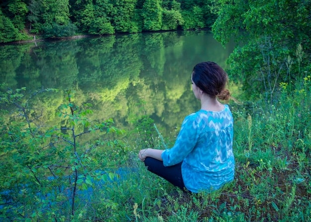 A person meditating in a serene environment, symbolizing mindfulness and self-reflection, with lush green trees and a gentle stream in the background.